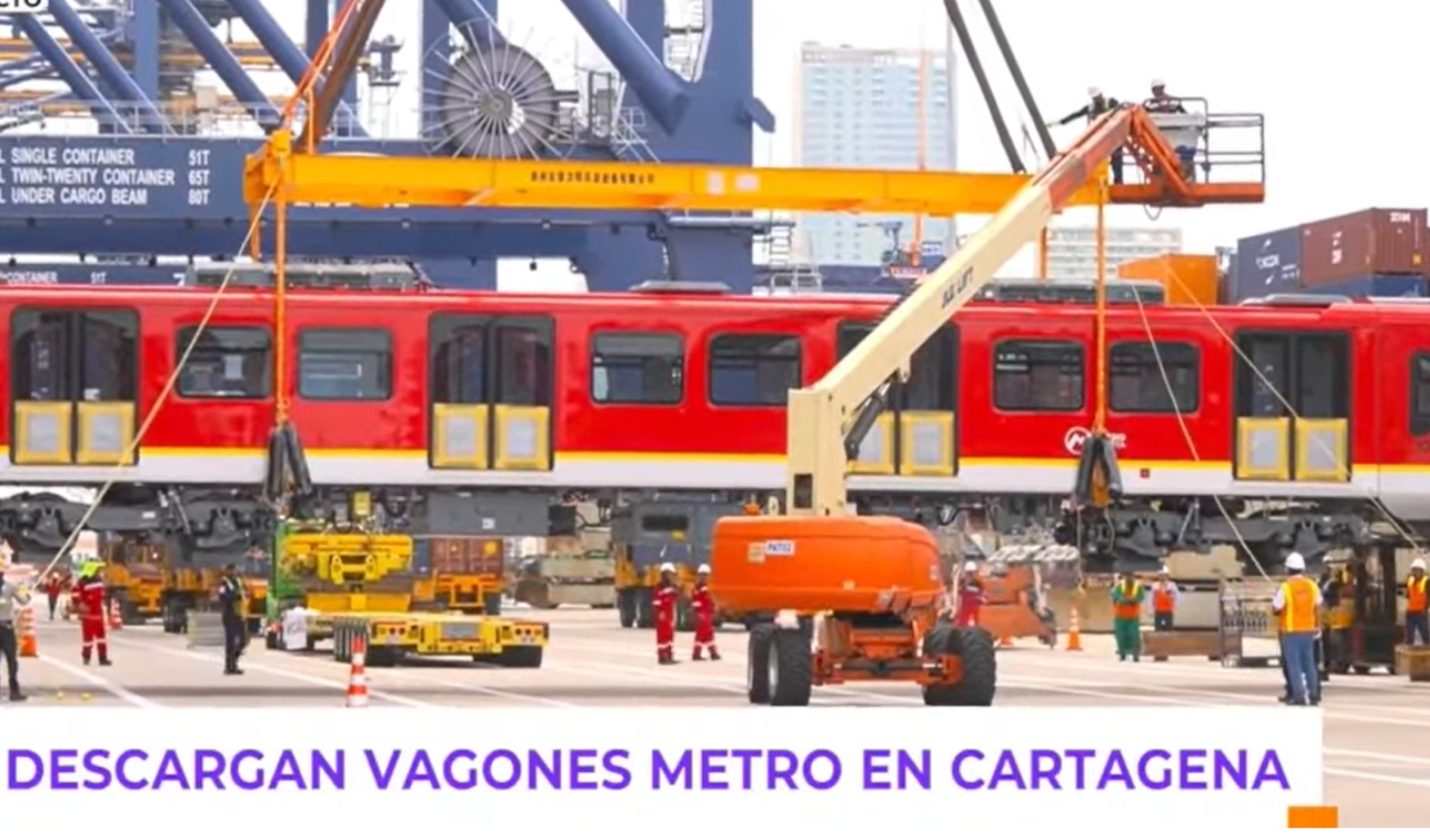 Vista lateral del tren mientras es bajado cuidadosamente del buque. Varias personas del equipo técnico coordinan la maniobra en tierra, con contenedores marítimos de fondo.